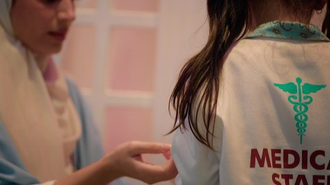 مفهوم التعليم عن طريق اللعب، تعريف الأطفال على المهن المستقبلية، أم إماراتية ترتدي عباءة تقوم باللعب مع ابنتها، الطب والرعاية الصحية، تصوير مقرب لفتاة عربية خليجية سعودية ترتدي الزي الطبي تقوم بمعالجة يد والدتها، الاستمتاع في مركز الالعاب الترفيهي