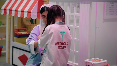 العمل في الطاقم الطبي، الاستمتاع في مركز الالعاب الترفيهي، مفهوم التعليم عن طريق اللعب، تعريف الأطفال على المهن المستقبلية، أم إماراتية ترتدي عباءة تقوم باللعب مع ابنتها، تصوير من الخلف لفتاة عربية خليجية سعودية ترتدي الزي الطبي تقوم بمعالجة يد والدتها