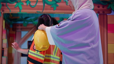 Playing in the work area at the Entertainment Games Center, Expressions of happiness and pleasure, an Arab -Emirati Gulf girl wearing helmet, photography from behind Arab Gulf Arab Saudi women dressing the protection jacket for her daughter, future professions and jobs, care and development of children's skills, spending fun times to play