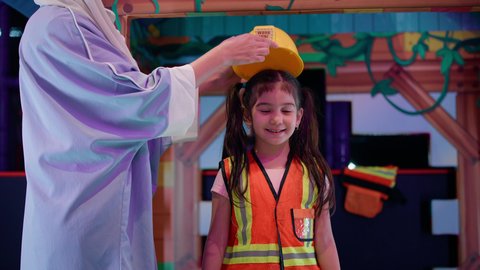 زتعابير السعادة والسرور، فتاة عربية خليجية إماراتية ترتدي سترة الحماية، تصوير مقرب لام عربية خليجية سعودية تقوم بوضع الخوذة الصفراء لابنتها، مهن ووظائف مستقبلية، رعاية وتطوير مهارات الأطفال، قضاء أوقات ممتعة باللعب، اللعب في منطقة العمل في مركز الالعاب الترفيهيه