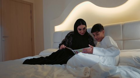 شغف قراءة القصص والروايات الممتع، مفهوم التثقيف والمعرفة، مفهوم التربيه القائمه على الحب والحنان، أم عربيه خليجيه اماراتية ترتدي العباءة  تستلقي على سرير مريح برفقة ولدها تقرأ الروايات المسليه