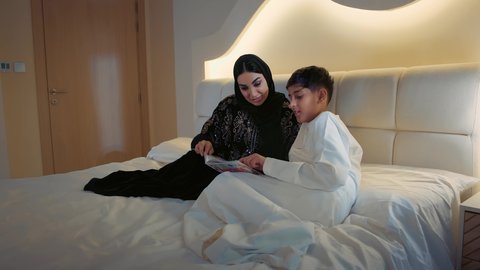 مفهوم التثقيف والمعرفة، المشاركه بقراءه القصص المفيده مع الأبناء قبل النوم، مفهوم التربيه القائمه على الحب والحنان، أم عربيه خليجيه اماراتية ترتدي العباءة  تستلقي على سرير مريح برفقة ولدها تقرأ الروايات المسليه