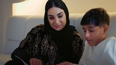 المشاركه بقراءه القصص المفيده مع الأبناء قبل النوم، مفهوم التربيه القائمه على الحب والحنان، تصوير مقرب لأم عربيه خليجيه اماراتية ترتدي العباءة  تستلقي على سرير مريح برفقة ولدها تقرأ الروايات المسليه