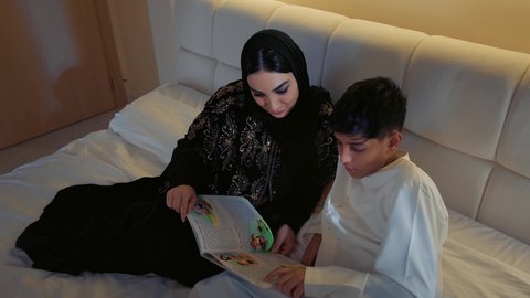 الإستمتاع بالأنشطه الترفيهية وقضاء الوقت مع الاولاد، قراءه القصص المفيده قبل النوم، مفهوم التربيه القائمه على الحب والحنان، أم عربيه خليجيه اماراتية ترتدي العباءة  تستلقي على سرير مريح برفقة ولدها تقرأ الروايات المسليه