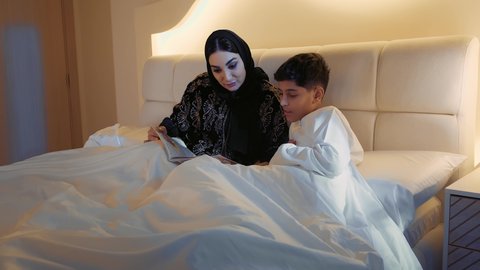 Encouraging children to learn and educate themselves, reading useful stories before bedtime, spending time with the son, the concept of parenting based on love and tenderness, a close-up of an Arab Gulf Emirati mother wearing an abaya lying on a comfortable bed with her son reading entertaining novels