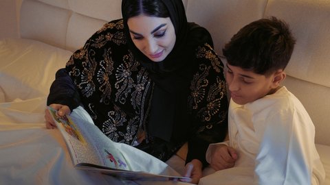 قراءه القصص المفيده قبل النوم، قضاء الوقت مع الابن، مفهوم التربيه القائمه على الحب والحنان، تصوير مقرب لأم عربيه خليجيه اماراتية ترتدي العباءة  تستلقي على سرير مريح برفقة ولدها تقرأ الروايات المسليه