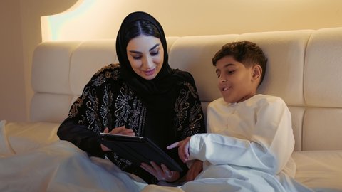 الثقة بحنان الأم ودورها في حياة الأبناء، استعمال الأجهزة التكنولوجية الحديثة والمتطورة, تصوير مقرب لام عربيه خليجيه اماراتية ترتدي الحجاب تجلس على السرير في غرفه النوم مع ابنها,ام سعودية تمسك بيدها جهاز التابليت المحمول