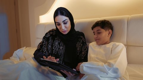 قضاء وقت ممتع مع الابن، استعمال الأجهزة التكنولوجية الحديثة والمتطورة, تصوير مقرب لام عربيه خليجيه اماراتية ترتدي الحجاب تجلس على السرير في غرفه النوم مع ابنها,ام سعودية تمسك بيدها جهاز التابليت المحمول
