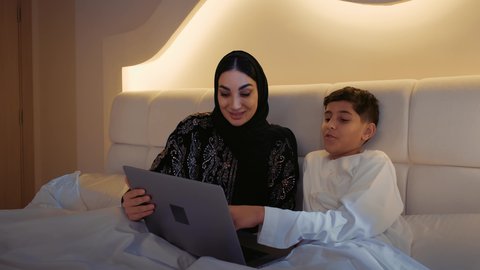 حل الواجبات المنزليه، دمج التعليم مع التقنية و التكنولوجيا، معلمة خصوصي لمتابعة الدروس في المنزل، أم عربية خليجة اماراتية محجبة تجلس على السرير تقوم بتعليم وتدريس ابنها باستخدام الحاسب المحمول، متابعة منصات التعليم الالكتروني المتاحة عبر الانترنت