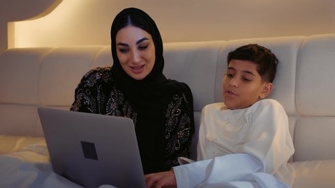 دمج التعليم مع التقنية و التكنولوجيا، حل الواجبات المنزليه، معلمة خصوصي لمتابعة الدروس في المنزل، أم عربية خليجة اماراتية محجبة تجلس على السرير تقوم بتعليم وتدريس ابنها باستخدام الحاسب المحمول، متابعة منصات التعليم الالكتروني المتاحة عبر الانترنت