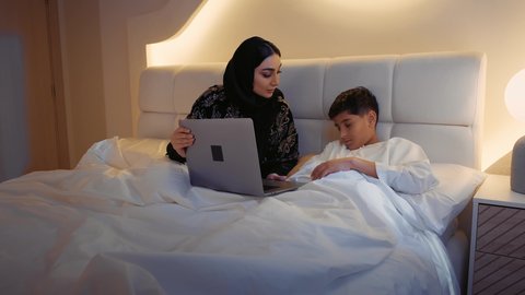 دور ومسؤوليه الأم تجاه أولادها، أجواء الراحة والهدوء، الاسترخاء والنوم بعمق،، قضاء الوقت مع الابن، سيدة سعودية محجبة تستلقي على سرير مريح برفقة ولدها،ام عربيه خليجيه اماراتية ترتدي العباءه تمسك جهاز الحاسب المحمول