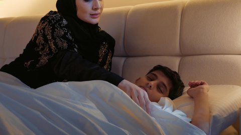 أجواء الراحة والهدوء، الاسترخاء والنوم بعمق، مفهوم العناية والاهتمام بالأولاد، قضاء الوقت مع الابن، سيدة سعودية محجبة تستلقي على سرير مريح برفقة ولدها،ام عربيه خليجيه اماراتية ترتدي العباءه تقوم بتغطية ابنها