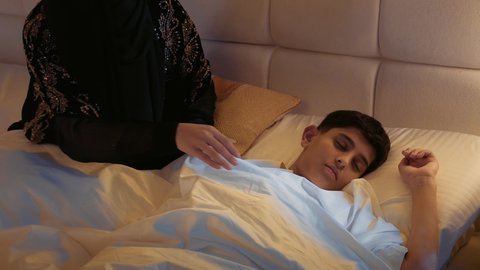 الاسترخاء والنوم بعمق، الشعور بالأمان والاطمئنان، مفهوم العناية والاهتمام بالأولاد، قضاء الوقت مع الابن، سيدة سعودية محجبة تستلقي على سرير مريح برفقة ولدها، تصوير مقرب لأم عربيه خليجيه اماراتية ترتدي العباءه تقوم بتغطية ابنها