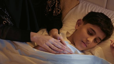 A feeling of love and care, the concept of motherhood and family warmth, spending time with the son, a veiled Saudi woman lying on a comfortable bed with her son, a close-up of the hand of an Arab Gulf Emirati mother wearing an abaya holding her son's hand and patting him