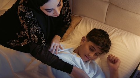 قضاء وقت ممتع مع الابناء، حنان الأمهات و تحمل المسؤولية والعناية بالأطفال، تصوير مقرب لام عربية خليجية اماراتية تجلس على السرير، سيده سعودية تقوم بتقبيل ابنها اثناء النوم بعطف وحب، مشاعر الأمومة والطفولة
