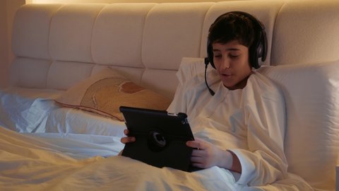 استخدام التكنولوجيا للتسلية، تصويرمقرب لطفل عربي اماراتي خليجي يرتدي الثوب التقليدي و الهيدفون يجلس على السرير ويقوم باستخدام التابلت، قضاء وقت ممتع على الإنترنت، اللعب على الجهاز اللوحي