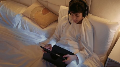 مفهوم التعليم وإنجاز المهام عن بعد، استخدام الأجهزة التقنية الحديثة، تصوير لطفل عربي اماراتي خليجي يرتدي الثوب التقليدي و الهيدفون يجلس على السرير ويقوم باستخدام التابلت، قضاء وقت ممتع على الإنترنت، اللعب على الجهاز اللوحي