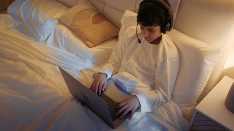 استخدام الأجهزة التقنية الحديثة المتطورة، قضاء وقت مريح وممتع في العطلة، الاسترخاء والاستلقاء في اوقات الفراغ، تصوير من الاعلى لولد عربي خليجي اماراتي يرتدي الدشداشة يستلقي على السرير في غرفة النوم يحمل الحاسب المحمول و يضع الهيدفون على راسه