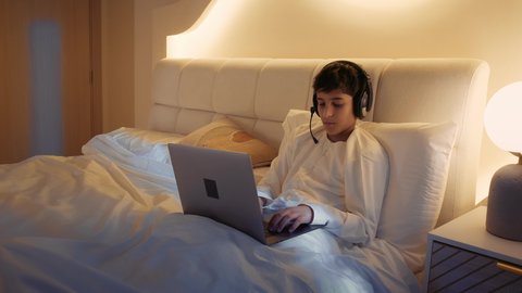 قضاء وقت مريح وممتع في العطلة، الاسترخاء والاستلقاء في اوقات الفراغ، استخدام الأجهزة التقنية الحديثة المتطورة، ولد عربي خليجي اماراتي يرتدي الدشداشة يستلقي على السرير في غرفة النوم يمسك الحاسب المحمول و يضع الهيدفون على راسه