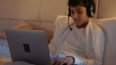 الاسترخاء والاستلقاء في اوقات الفراغ، قضاء وقت مريح وممتع في العطلة، استخدام الأجهزة التقنية الحديثة المتطورة، ، تصوير مقرب لولد عربي خليجي اماراتي يرتدي الدشداشة يستلقي على السرير في غرفة النوم يمسك الحاسب المحمول و يضع الهيدفون على راسه