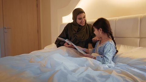 انشطة ترفيهية قبل النوم،  قراءة القصص والروايات، اهتمام الأم بالابناء، مفهوم السعادة الاسرية، تصويرمقرب لامرأة عربية خليجية اماراتية تجلس على السرير تقرأ لإبنتها قصه مسلية، ام سعودية تقضي وقت ممتع مع ابنتها
