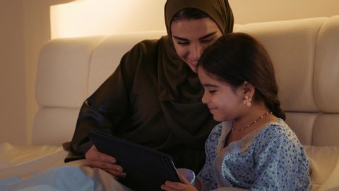 استخدام الاجهزه الحديثة و التقنية، تصويرمقرب لسيدة عربيه خليجيه سعودية ترتدي العباءه الملونه تجلس مع طفلتها على السرير ،ام اماراتية تقوم بلعب مع طفلتها بجهاز التابلت
