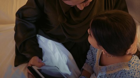 Spending beautiful times with children, a close-up of a Saudi Arabian Gulf woman wearing a colorful abaya sitting with her daughter on the bed, an Emirati mother playing with her daughter on a tablet, using modern devices and technology