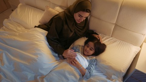 المحافظة على هدوء الطفلة، الاسترخاء والشعور بالأمان، سيدة سعودية تقوم بمداعبة الطفلة بلطف وحنان، صورة مقربة لبنت عربية خليجية اماراتية تستلقي في السرير تمسك والدتها بيدها، ملامح ناعمة وبريئة