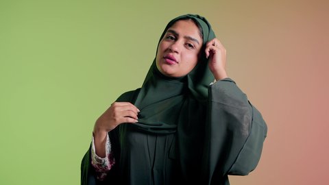 ترتيب الحجاب والاستعداد للذهاب، ارتداء عباءة ملونة عصرية، الإهتمام بالموضة والجمال والأناقة، تصوير مقرب لإمرأة عربية سعودية خليجية تنظر إلى الكاميرا بإيماءات الثقة بالنفس، عرض الملابس والاكسسوارات النسائية السعودية، خلفية ملونة