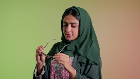 الأناقة والاهتمام بالمظهر الخارجي، سيدة اماراتية تقوم بالعروض الإعلانية التسويقية للمنتجات المحلية، عرض احدث موديلات النظارات الشمسية، اتباع آخر صيحات الموضة, تصوير مقرب لإمرأة عربية خليجية سعودية مبتسمة ترتدي الحجاب والعباءة تقوم بوضع النظارة، خلفية ملونة