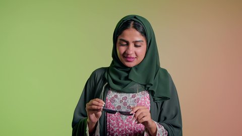 العروض الإعلانية التسويقية للمنتجات المحلية، الأناقة والاهتمام بالمظهر الخارجي، سيدة اماراتية تقوم بعرض احدث موديلات النظارات الشمسية، اتباع آخر صيحات الموضة, تصوير مقرب لإمرأة عربية خليجية سعودية مبتسمة ترتدي الحجاب والعباءة تقوم بوضع النظارة، خلفية ملونة
