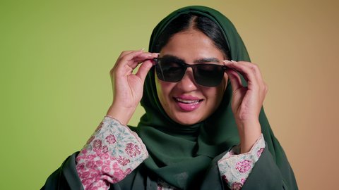 التسويق للمنتجات المحلية، الأناقة والاهتمام بالمظهر الخارجي، سيدة اماراتية تقوم بعرض احدث موديلات النظارات الشمسية، اتباع آخر صيحات الموضة, تصوير مقرب لإمرأة عربية خليجية سعودية مبتسمة ترتدي الحجاب والعباءة تقوم بوضع النظارة، خلفية ملونة