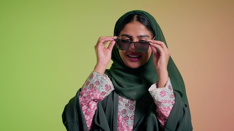 ارتداء النظارة والاهتمام بالمظهر الخارجي والاناقة الشخصية، سيدة اماراتية تقوم بعرض احدث موديلات النظارات الشمسية، متابعة آخر صيحات الموضة, تصوير مقرب لإمرأة عربية خليجية سعودية مبتسمة ترتدي الحجاب والعباءة تقوم بالتسويق للمنتجات المحلية، خلفية ملونة
