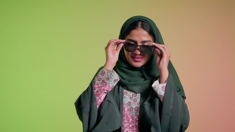 العروض الإعلانية التسويقية للمنتجات المحلية، الأناقة والاهتمام بالمظهر الخارجي، سيدة اماراتية تقوم بعرض احدث موديلات النظارات الشمسية، اتباع آخر صيحات الموضة, تصوير مقرب لإمرأة عربية خليجية سعودية مبتسمة ترتدي الحجاب والعباءة تقوم بوضع النظارة، خلفية ملونة
