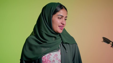 ايماءات الانوثة والسرور، ارتداء النظارة والاهتمام بالمظهر الخارجي والاناقة الشخصية، سيدة اماراتية تقوم بعرض احدث موديلات النظارات الشمسية، تصوير مقرب لإمرأة عربية خليجية سعودية مبتسمة ترتدي الحجاب والعباءة تقوم بالتسويق للمنتجات المحلية، خلفية ملونة