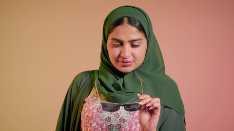 الأناقة والاهتمام بالمظهر الخارجي، سيدة اماراتية تقوم بالعروض الإعلانية التسويقية للمنتجات المحلية، عرض احدث موديلات النظارات الشمسية، اتباع آخر صيحات الموضة, تصوير مقرب لإمرأة عربية خليجية سعودية مبتسمة ترتدي الحجاب والعباءة تقوم بوضع النظارة، خلفية ملونة