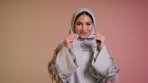 Following the latest fashion trends, close-up of a smiling Saudi Arabian Gulf woman wearing a hijab and abaya putting on glasses, elegance and interest in outward appearance, an Emirati woman doing marketing advertisements for local products, displaying the latest models of sunglasses, colorful background