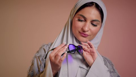 ارتداء النظارة والاهتمام بالمظهر الخارجي والاناقة الشخصية، سيدة اماراتية تقوم بعرض احدث موديلات النظارات الشمسية، متابعة آخر صيحات الموضة, تصوير مقرب لإمرأة عربية خليجية سعودية مبتسمة ترتدي الحجاب والعباءة تقوم بالتسويق للمنتجات المحلية، خلفية ملونة