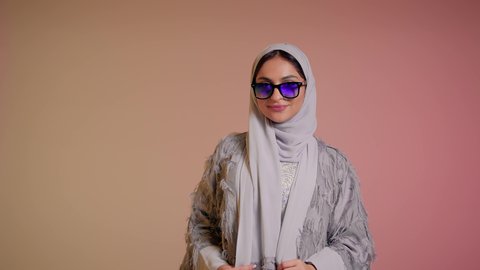 ايماءات الانوثة والسرور، ارتداء النظارة والاهتمام بالمظهر الخارجي والاناقة الشخصية، سيدة اماراتية تقوم بعرض احدث موديلات النظارات الشمسية، تصوير مقرب لإمرأة عربية خليجية سعودية مبتسمة ترتدي الحجاب والعباءة تقوم بالتسويق للمنتجات المحلية، خلفية ملونة