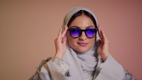 ارتداء النظارة والاهتمام بالمظهر الخارجي والاناقة الشخصية، سيدة اماراتية تقوم بعرض احدث موديلات النظارات الشمسية، متابعة آخر صيحات الموضة, تصوير مقرب لإمرأة عربية خليجية سعودية مبتسمة ترتدي الحجاب والعباءة تقوم بالتسويق للمنتجات المحلية، خلفية ملونة