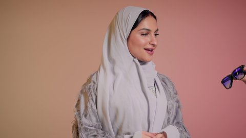 تعابير الدهشة والسرور، ارتداء النظارة والاهتمام بالمظهر الخارجي والاناقة الشخصية، سيدة اماراتية تقوم بعرض احدث موديلات النظارات الشمسية، تصوير مقرب لإمرأة عربية خليجية سعودية مبتسمة ترتدي الحجاب والعباءة تقوم بالتسويق للمنتجات المحلية، خلفية ملونة