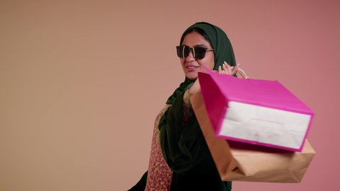 ايماءات السعادة والسرور، الشراء من الاسواق والمتاجر، الاُثر الايجابي للتسوق، شابة امارتية تستفيد من العروض والتخفيض الموسمي، تصوير مقرب لامرأة عربية خليجية سعودية ترتدي العباءة والحجاب تحمل بيديها الاكياس، اقتناء الهدايا والمستلزمات، خلفية ملونة