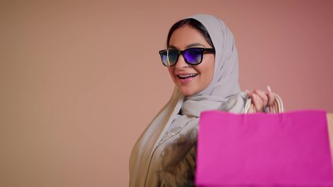Positive impact of shopping, young Emirati woman holding bags with gestures of pleasure, taking advantage of seasonal offers and discounts, close-up of a Saudi Arabian Gulf woman wearing an abaya and hijab holding bags, buying gifts and supplies, shopping from markets and stores, colorful background