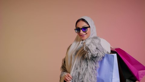 شابة اماراتية تمسك الحقائب بإيماءات السرور، الاُثر الايجابي للتسوق، الاستفادة من العروض والتخفيض الموسمي، تصوير مقرب لامرأة عربية خليجية سعودية ترتدي العباءة والحجاب تحمل بيديها الاكياس، اقتناء الهدايا والمستلزمات، الشراء من الاسواق والمتاجر، خلفية ملونة