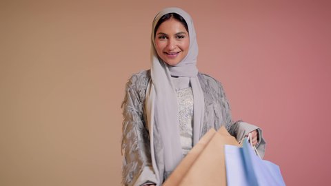 شراء الهدايا والمستلزمات، الاستفادة من العروض والتخفيض الموسمي، الاُثر الايجابي للتسوق على نفسية النساء، تصوير مقرب لامرأة عربية خليجية سعودية ترتدي العباءة والحجاب تحمل بيديها أكياس ملونة، سيدة اماراتية تمسك الحقائب بإيماءات السرور، خلفية ملونة