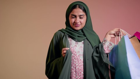 شراء الهدايا والمستلزمات، الاستفادة من العروض والتخفيض الموسمي، الاُثر الايجابي للتسوق على نفسية النساء، تصوير مقرب لامرأة عربية خليجية سعودية ترتدي العباءة والحجاب تحمل بيديها أكياس ملونة، سيدة اماراتية تمسك الحقائب بإيماءات السرور، خلفية ملونة