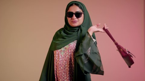 شراء بضائع الماركات العالمية، الاهتمام بالمظهر الخارجي والاناقة، متابعة آخر صيحات الموضة, تصوير مقرب لامرأة عربية خليجية اماراتية ترتدي الحجاب والعباءة مع النظارات الشمسية تقف باستقامة بتعابير الثقة بالنفس، سيدة سعودية تنظر الى الكاميرا وتحمل بيدها حقيبة حمراء، خلفية ملونة