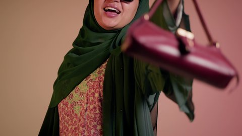 الاهتمام بالمظهر الخارجي والاناقة، شراء بضائع الماركات العالمية، متابعة آخر صيحات الموضة, تصوير مقرب لامرأة عربية خليجية اماراتية ترتدي الحجاب والعباءة مع النظارات الشمسية تقف باستقامة بتعابير الثقة بالنفس، سيدة سعودية تنظر الى الكاميرا وتحمل بيدها حقيبة حمراء، خلفية ملونة