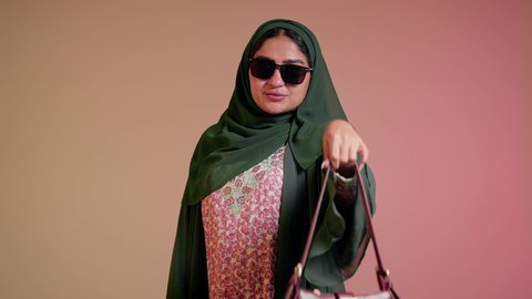 متابعة آخر صيحات الموضة, شراء بضائع الماركات العالمية، الاهتمام بالمظهر الخارجي والاناقة، تصوير مقرب لامرأة عربية خليجية اماراتية ترتدي الحجاب والعباءة مع النظارات الشمسية تقف باستقامة بتعابير الثقة بالنفس، سيدة سعودية تنظر الى الكاميرا وتحمل بيدها حقيبة حمراء، خلفية ملونة
