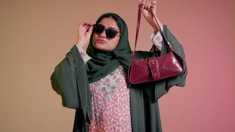 Caring about appearance and elegance, buying international brand goods, following the latest fashion trends, close-up of an Emirati Gulf Arab woman wearing a hijab and abaya with sunglasses, standing straight with an expression of self-confidence, a Saudi woman looking at the camera and holding a red bag, colorful background